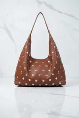 Bolso tipo hobo de cuero marrón, adornado con tachas metálicas redondas plateadas en toda su superficie. Posee un asa de hombro única y delgada.