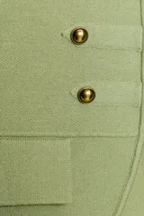Chaleco de punto en color verde claro, con cuello redondo y diseño sin mangas. Presenta un estilo militar con detalles de alamares, botones decorativos dorados en el frente y bolsillos falsos con tapeta. Cierre frontal con corchetes ocultos.