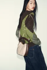 Bolso tipo saca con diseño estructurado, confeccionado en una combinación de tejido de lana en tono beige y laterales de cuero sintético marrón. Presenta costuras decorativas verticales, asa de mano superior y una correa bandolera extraíble con detalle de franjas. Cuenta con cierre mediante botón imantado.