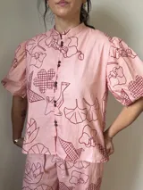 Camisa de lino y algodón color rosado, con estampado de motivos lineales en tono bordó, cuello mao, cierre frontal con botones agrupados y mangas cortas abullonadas.