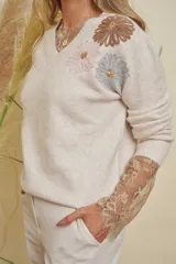 Buzo color beige con cuello en V y mangas largas. Presenta apliques de flores bordadas en tonos marrón, rosa y celeste, con lentejuelas doradas en el centro. Las mangas tienen un detalle de encaje beige que sobresale de los puños.