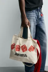 Bolso tipo shopper de tela color beige, con asas cortas y asa de hombro. Estampado frontal con cuatro dados rojos con corazones y texto que dice "SOMETIMES SÍ, SOMETIMES BYE".