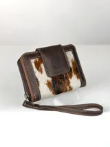 Mini billetera de cuero marrón con detalles en cuero con pelo con estampado animal print tipo vaca en tonos blancos, marrones y negros. Cuenta con solapa frontal con botón a presión y diseño de búho grabado, ribetes pespunteados y correa de mano.