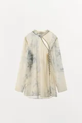Camisa de cuello redondo y manga larga con diseño de estampado artístico en tonos azules sobre fondo beige. Presenta un cierre frontal asimétrico con botones forrados a tono y una abertura lateral en el escote.