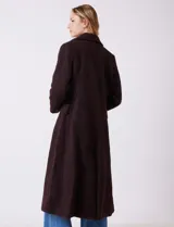 Tapado largo de bouclé color marrón oscuro, con silueta entallada que termina en evasé. Tiene cuello de solapa amplia, cierre frontal con tres botones a tono y bolsillos funcionales con solapa. Incluye forro interno a tono.