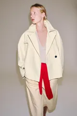 Trench corto color marfil de algodón, con cuello camisero, solapa cortaventos, mangas largas amplias con puños abotonados y cierre cruzado con un botón visible.