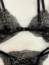 Conjunto de lencería compuesto por bralette triangular y tanga, confeccionado en encaje bitono negro y blanco. El bralette presenta cierre frontal con broche de gancho y breteles regulables. La tanga es de estilo colaless con laterales regulables mediante herrajes metálicos.