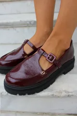 Zapato tipo guillermina en cuero charol color bordó, con suela track negra de goma, cierre con hebilla ajustable y detalle de herrajes metálicos dorados.