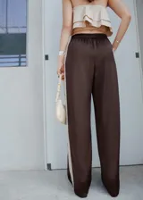 Pantalón de corte recto y tiro medio, confeccionado en tejido satinado color marrón oscuro. Presenta cintura con elástico, bolsillos laterales y una sutil raya lateral en contraste.