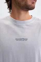 Remera de manga corta en color gris melange con efecto lavado. Presenta una estampa gráfica en la espalda con el texto 'Uncover Places' y detalles tipográficos en tonos gris oscuro y azul.