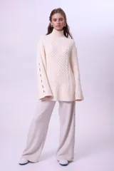 Sweater de punto color crema, con cuello alto y diseño de trenzas en el frente. Presenta mangas largas con aberturas laterales y corte holgado.