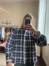 Camisa de corte holgado con estampado de cuadros en tonos azul, rojo y blanco. Presenta cuello alto, cierre frontal con botones y detalles de puntilla blanca en la tapeta central y en los puños abullonados.