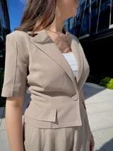 Blazer corto color beige, de manga corta, con solapas y cierre frontal con un botón. Presenta bolsillos de parche en la parte delantera.
