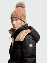 Gorro de lana de punto acanalado en color tostado, con un pompón de pelo en la parte superior y el parche con el logo de Moncler bordado en el frente del doblez.
