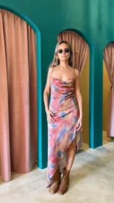 Vestido largo de crepé con estampado floral en tonos rosados, morados y blancos. Presenta escote buche con drapeado y espalda baja ajustable con tiras finas. El ruedo es irregular, más corto en el frente.