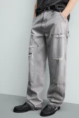 Pantalón de jean estilo carpintero con corte relaxed fit, tiro medio y pierna recta. Presenta un lavado azul medio con efecto desgastado y roturas en las rodillas. Incluye bolsillos laterales tipo plastrón, tira multifunción en la pierna, cierre frontal con cremallera y botón.