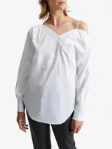 Camisa blanca de algodón con diseño asimétrico, escote cruzado con botones en diagonal, hombro descubierto con tirante fino, manga larga con puños abotonados y bolsillo de parche en el pecho.