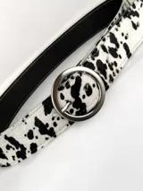 Cinturón de cuero genuino con estampado animal print tipo vaquita en blanco y negro. Presenta una hebilla circular metálica plateada.