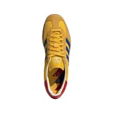 Championes Adidas Gazelle Indoor de diseño clásico, con exterior en tono amarillo con estampado geométrico sutil, tres bandas laterales en azul y detalle en rojo en el talón. Presenta puntera de gamuza y suela de caucho color caramelo.