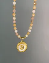 Collar de cristales facetados en tonos rosados y dorados, con dije circular de nácar y letra dorada en relieve.