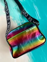 Bandolera Kaira de la línea Ottenta de Carolina De Cunto, confeccionada en material metalizado con acabado multicolor y efecto tornasolado. Cuenta con un bolsillo frontal con broche imantado, cierre superior y una correa larga y ajustable de tela negra con el nombre de la marca en letras blancas.