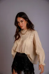 Camisa color beige de manga larga con cuello alto fruncido y detalles de encaje y pliegues en el canesú. Corte holgado.