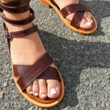 Sandalias planas de cuero color marrón, con tiras cruzadas en el empeine y múltiples tiras que se enrollan en la pierna, ajustables con hebillas metálicas.