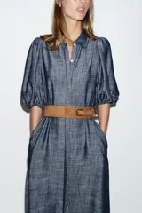Vestido midi de denim azul con cuello camisero, mangas abullonadas y cierre frontal oculto con botones. Incluye un cinturón marrón con hebilla metálica.