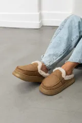 Zueco de gamuza color beige, con interior forrado en corderito sintético, suela de goma y detalle de hebilla lateral.