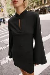 Vestido corto negro de cuello mao y escote en V, con mangas largas abanicadas y cierre de alamares en el frente. Cuenta con bolsillos frontales de plastrón y diseño de inspiración oriental.