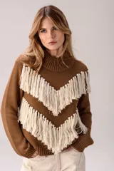 Sweater de punto color crema con cuello medio alto y silueta relajada. Presenta un diseño frontal con líneas en V decoradas con flecos largos de color marrón aplicados a mano.