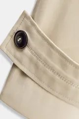 Trench corto color verde militar, con cuello solapa y capucha. Tiene manga larga con trabillas y botón en los puños, bolsillos delanteros y bajo asimétrico. Cierre frontal con botones. El tejido es repelente al agua.