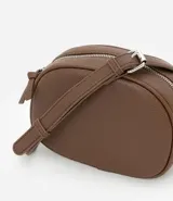 Cartera tipo camera bag pequeña, color marrón camel, de forma ovalada con superficie texturizada. Cuenta con correa ajustable larga, cierre principal con cremallera dorada y un pequeño bolsillo frontal con cremallera.