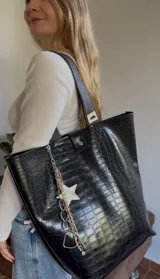 Bolso tipo tote bag negro con textura símil cuero de cocodrilo, doble asa corta marrón y correa larga al hombro. Incluye charm colgante con cadena plateada y estrella blanca.