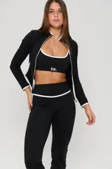 Conjunto deportivo de tres piezas compuesto por un top con breteles finos, una campera entallada con cierre frontal y una calza semioxford. Todas las prendas son de color negro con detalles de ribetes blancos en los bordes.