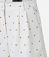 Bermuda de piquet color blanco con estampado de lunares negros, de corte recto, tiro alto, con trabillas en la cintura, pliegues y cierre tradicional.