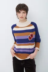 Polera de tejido de punto con diseño de rayas horizontales en tonos azul, marrón, crema y lila. Presenta cuello alto, mangas largas y un aplique bordado de flores rojas en el pecho.