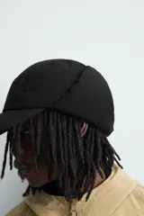 Gorra técnica de color negro con diseño de cinco paneles y orejeras integradas ajustables mediante una trabilla superior. Confeccionada en tejido acolchado, cuenta con ajuste elástico en la parte posterior.