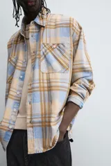 Camisa de manga larga con cuello solapa y dos bolsillos frontales de plastrón con botón. Presenta un estampado de cuadros en tonos beige, azul claro y marrón.