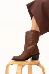 Bota estilo texana de caña media, confeccionada en cuero color marrón con acabado texturizado. Presenta un diseño clásico con costuras reforzadas, tiradores laterales y un taco cuadrado de altura media en color negro.