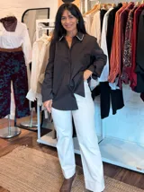 Camisa blanca de corte clásico y diseño holgado, con cuello camisero que presenta un detalle de pedrería o brillo sutil en los bordes. Posee mangas largas con puños rectos y un largo asimétrico, siendo más corta en la parte delantera y más larga en la parte trasera.