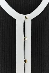 Chaqueta de punto acanalado en color negro con detalles en blanco en cuello, tapeta de botones, bolsillos y volados en el ruedo y puños. Presenta cierre frontal con botones dorados.