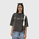 Remera de corte oversize con efecto acid wash en color gris oscuro. Presenta estampado gráfico en color amarillo en el frente con el logo de la marca y detalles de texto.