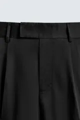 Pantalón negro de corte relaxed fit y estilo wide leg, con pliegues frontales dobles, bolsillos delanteros y detalle de bolsillos traseros de vivo. Cierre frontal cruzado con cremallera y gancho metálico oculto.