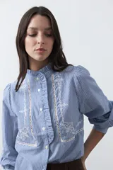 Camisa de rayas verticales con cuello mao, detalle de volados en el escote, bordados florales y recortes con calados. Posee mangas largas abullonadas con puños ajustados y cierre frontal con botones.