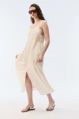 Vestido largo y amplio de color blanco, con escote cuadrado con detalles de puntilla, breteles finos ajustables con nudo, y fruncidos elásticos en la parte superior. Cuenta con una abertura frontal abotonada, bolsillos laterales y forro interno de voile de algodón.