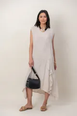 Vestido midi de lino color beige, sin mangas y con cuello redondo. Cuenta con un diseño irregular con detalle de volado vertical a un costado.
