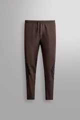 Pantalón de entrenamiento slim fit marrón oscuro de Zara Athleticz, confeccionado en tejido técnico elástico con paneles negros a contraste en las pantorrillas. Cuenta con cintura elástica ajustable con cordón.