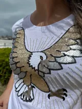 Remera blanca de algodón con cuello redondo y manga corta. Presenta un bordado de un águila con lentejuelas doradas, plateadas y negras en el frente.