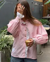 Camisa de manga larga abullonada en color rosa con rayas verticales finas. Presenta un diseño con volados en el cuello y a lo largo de la botonadura frontal, puños con detalle de rayas contrastantes y botones decorativos dorados.